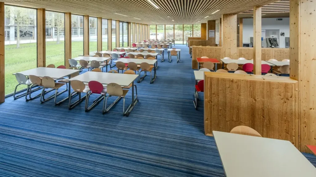 Spacious school cafeteria featuring blue striped carpet flooring, wooden pillars, and dining tables with colorful chairs.